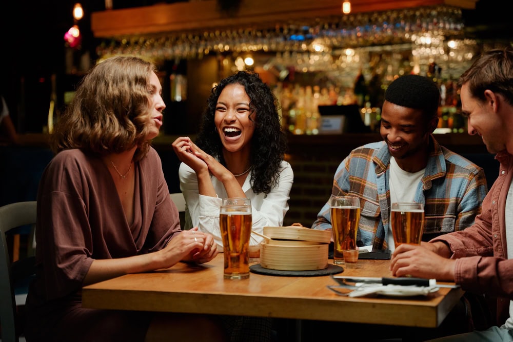 Four people sit at a table in a bar or restaurant, laughing and talking, with glasses of beer and food on the table in front of them.