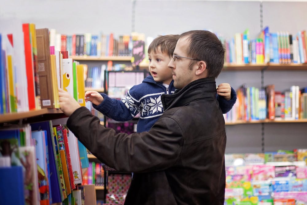 An adult holding a child browses books on a shelf in a bookstore, with colorful book covers and shelves visible in the background.
