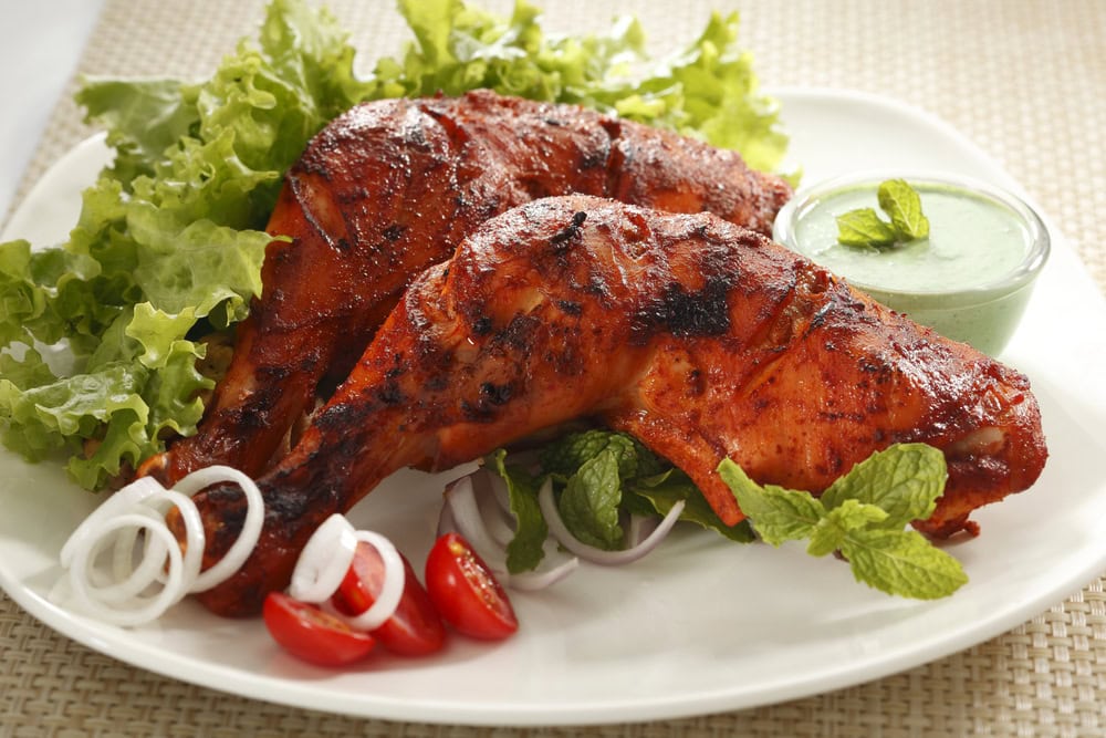 Grilled tandoori chicken legs served on a white plate with lettuce, sliced onions, cherry tomatoes, mint leaves, and a small bowl of green dip.