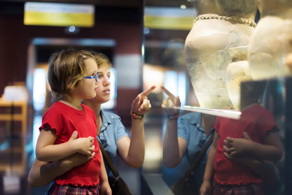 An adult and a child stand in front of a glass display case at a museum, looking at and pointing toward a large ceramic artifact.