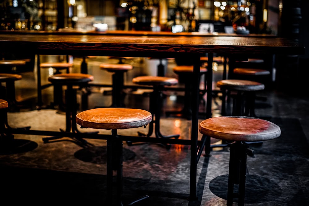 A group of round, cushioned bar stools are arranged around a high wooden table in a dimly lit bar or restaurant interior.