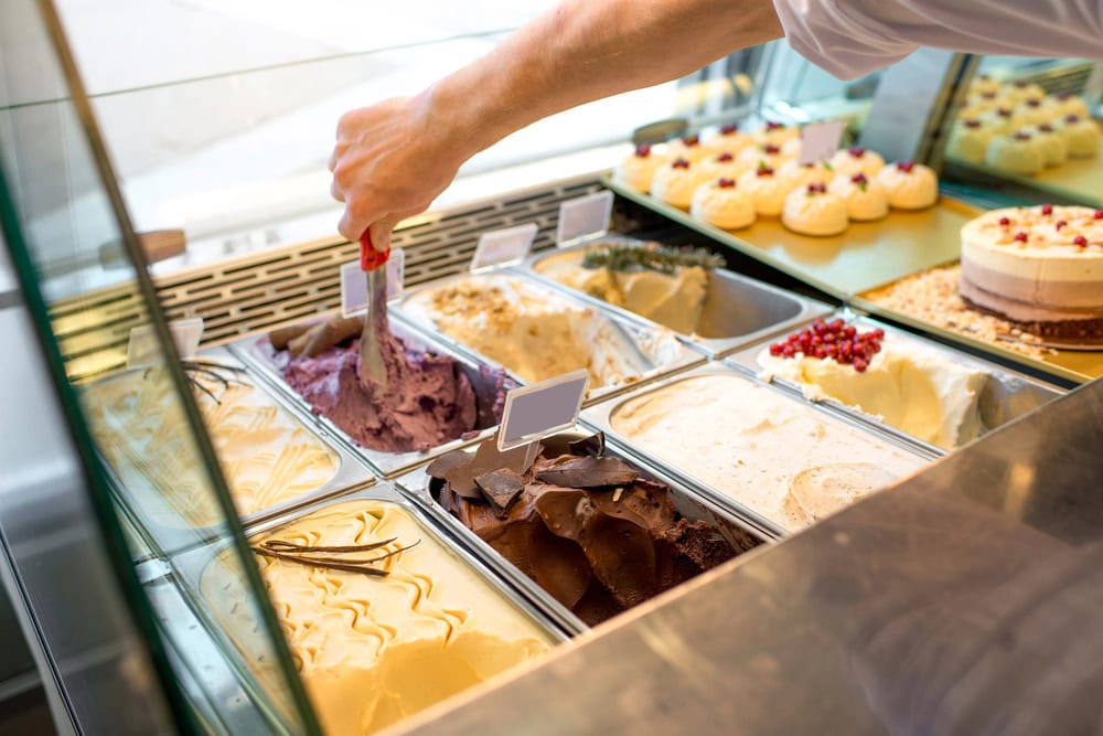 A person scoops purple gelato from a display case filled with various gelato flavors; desserts and cakes are visible in the background.