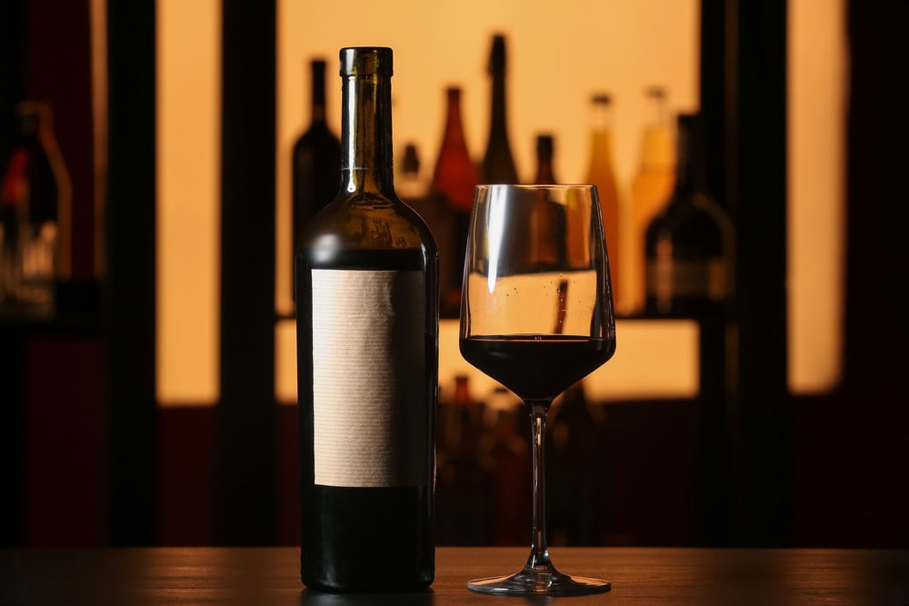 A bottle of red wine with a blank label stands next to a filled wine glass on a dark surface, with blurred bottles in the background.