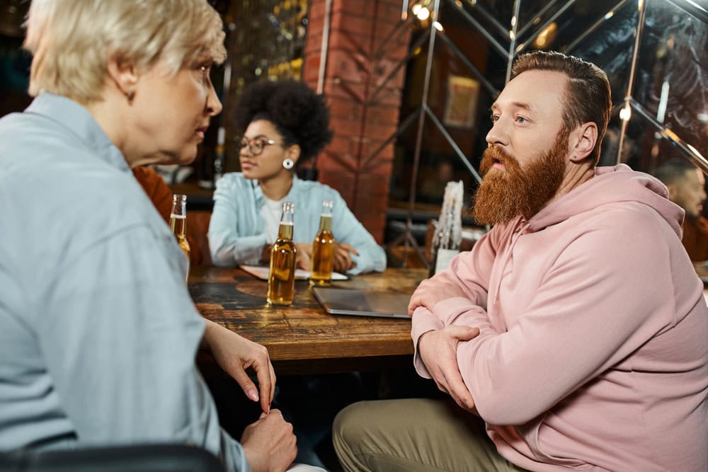 Four people sit at a table in a bar or restaurant, engaged in conversation, with beer bottles on the table.