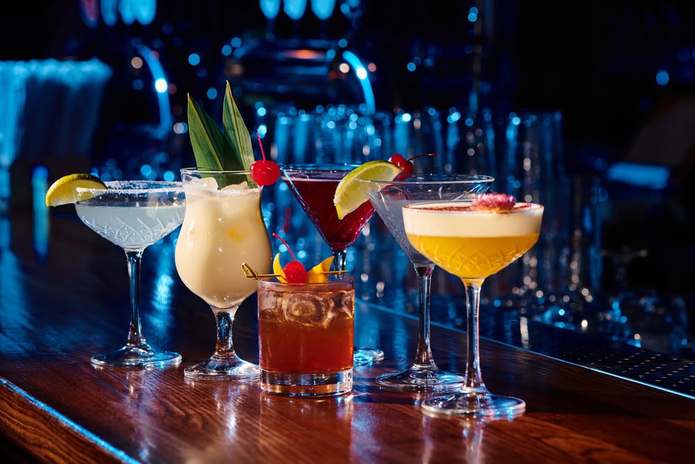 Five colorful cocktails in different glassware are arranged on a wooden bar top, with a blurred bar background and blue lighting.