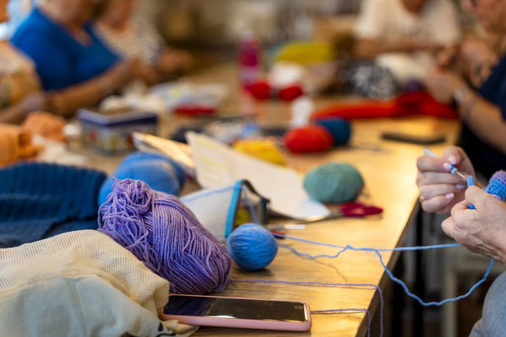 People are sitting around a table working with colorful yarn, knitting or crocheting, with supplies and a smartphone on the table.