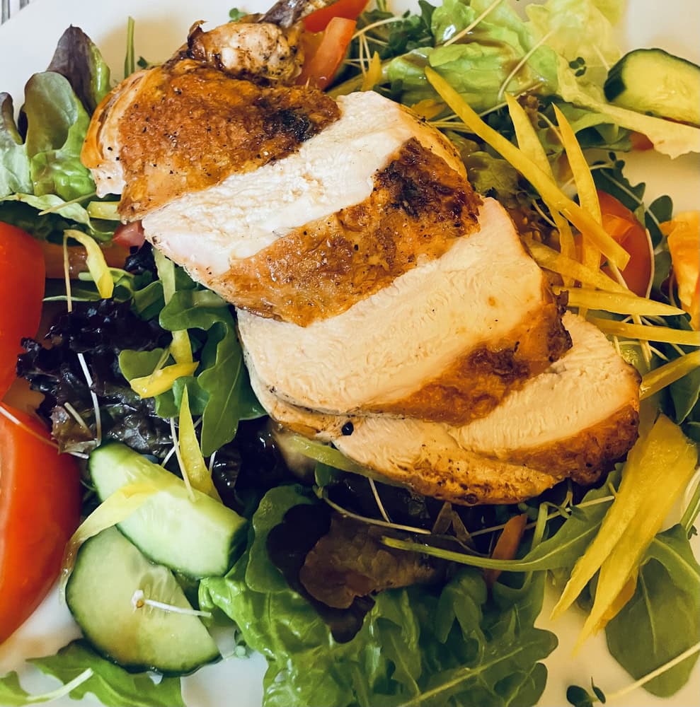 Sliced grilled chicken breast served on a bed of mixed salad greens, with tomato, cucumber, and yellow bell pepper strips.