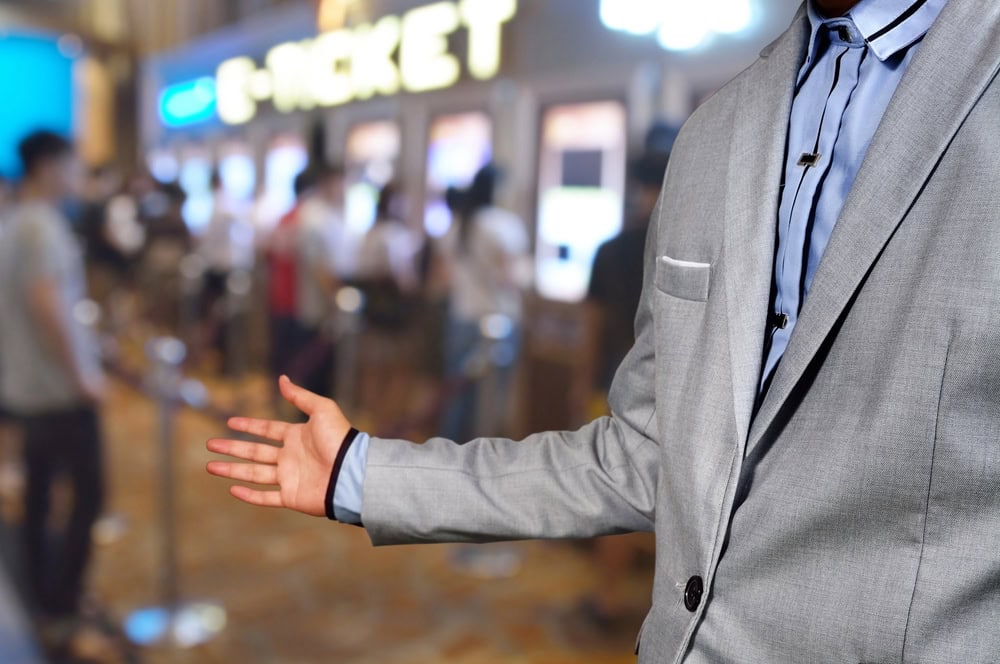 A person in a gray suit gestures toward a ticket counter with people waiting in line in the background.