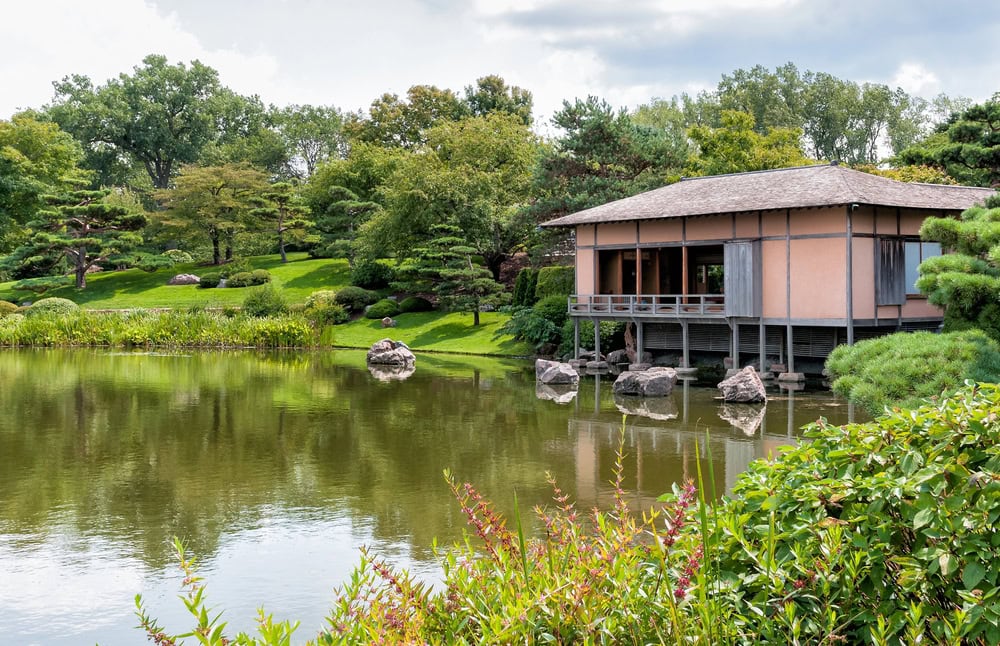 A traditional Japanese tea house stands over a calm pond surrounded by green trees and manicured shrubs in a serene garden setting.