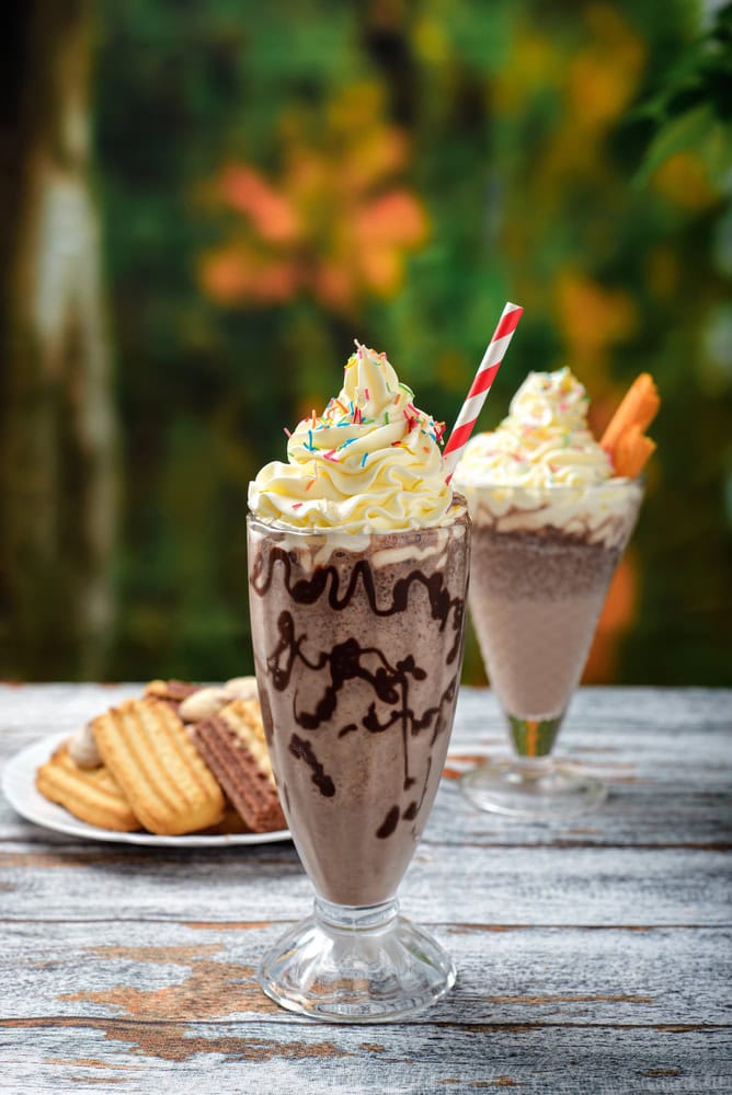 Two tall milkshakes with whipped cream and sprinkles, served in glasses with straws, sit on a wooden table; plate of assorted cookies in the background.