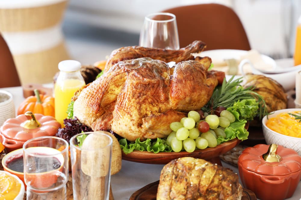 A roast turkey on a platter with grapes and lettuce is surrounded by various side dishes, drinks, and small pumpkins on a festive dining table.