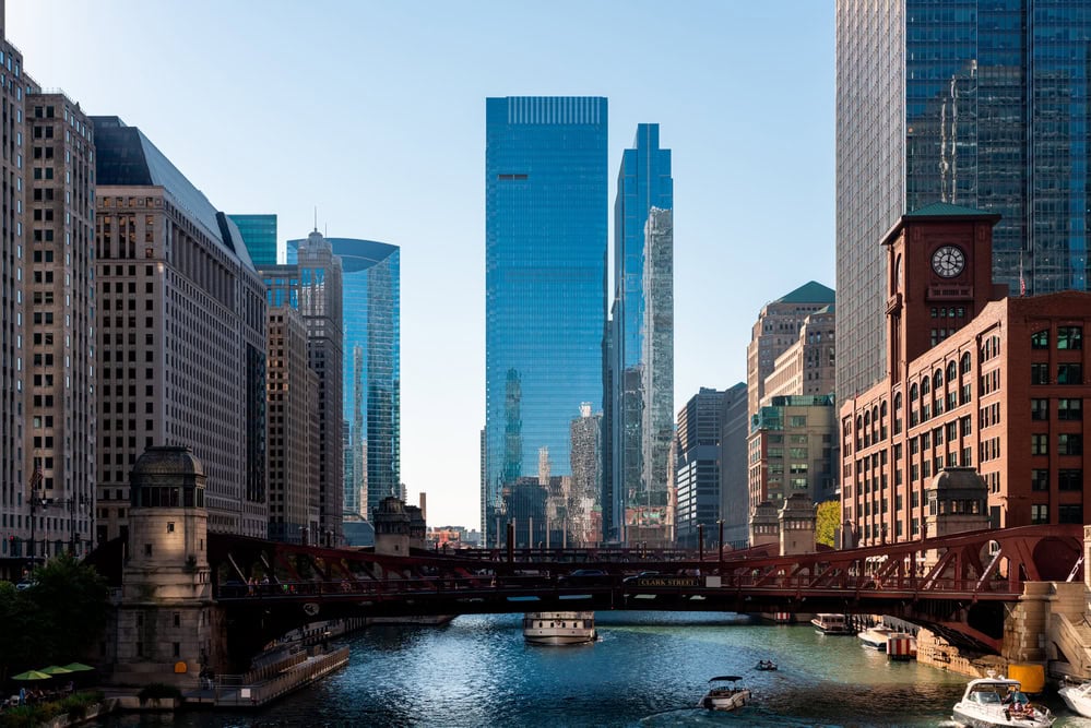 A river runs through a city with modern glass skyscrapers and historic brick buildings on either side, with boats visible on the water and a bridge in the foreground.