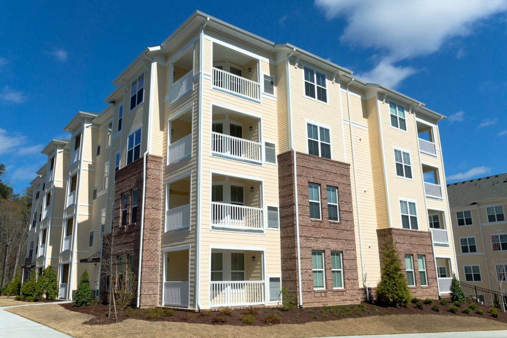Four-story apartment building with light yellow siding, brick accents, white balconies, and multiple windows under a partly cloudy sky.