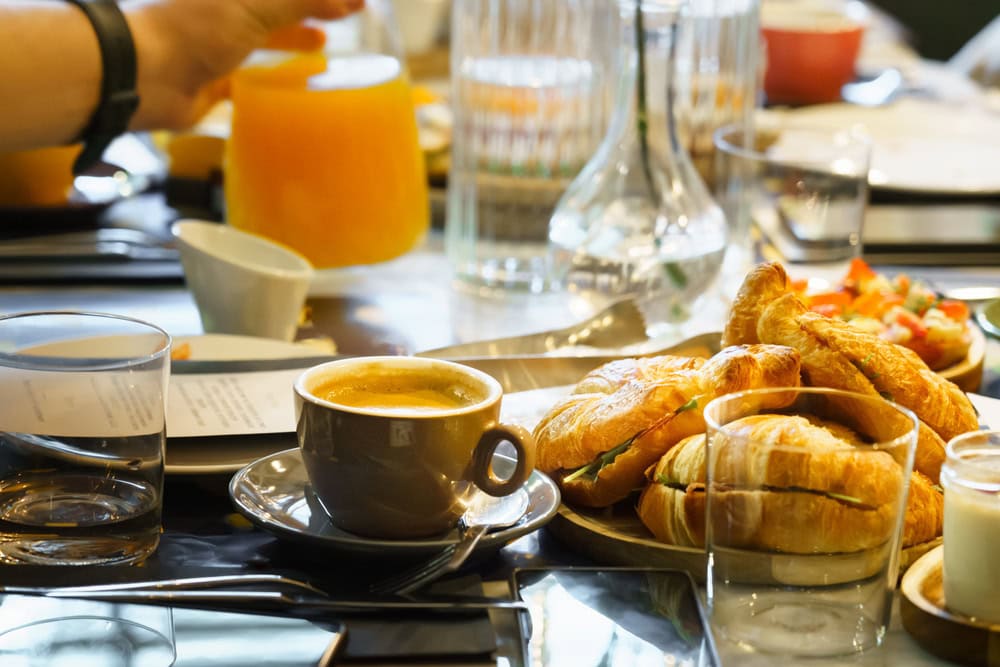 A breakfast table with a cup of coffee, a glass of water, a plate of croissants, and someone pouring orange juice in the background.