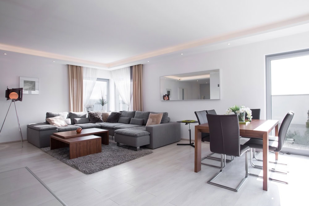 Modern living and dining area with a gray sectional sofa, wooden coffee table, dining table with six chairs, large mirror, and floor-to-ceiling windows with light curtains.