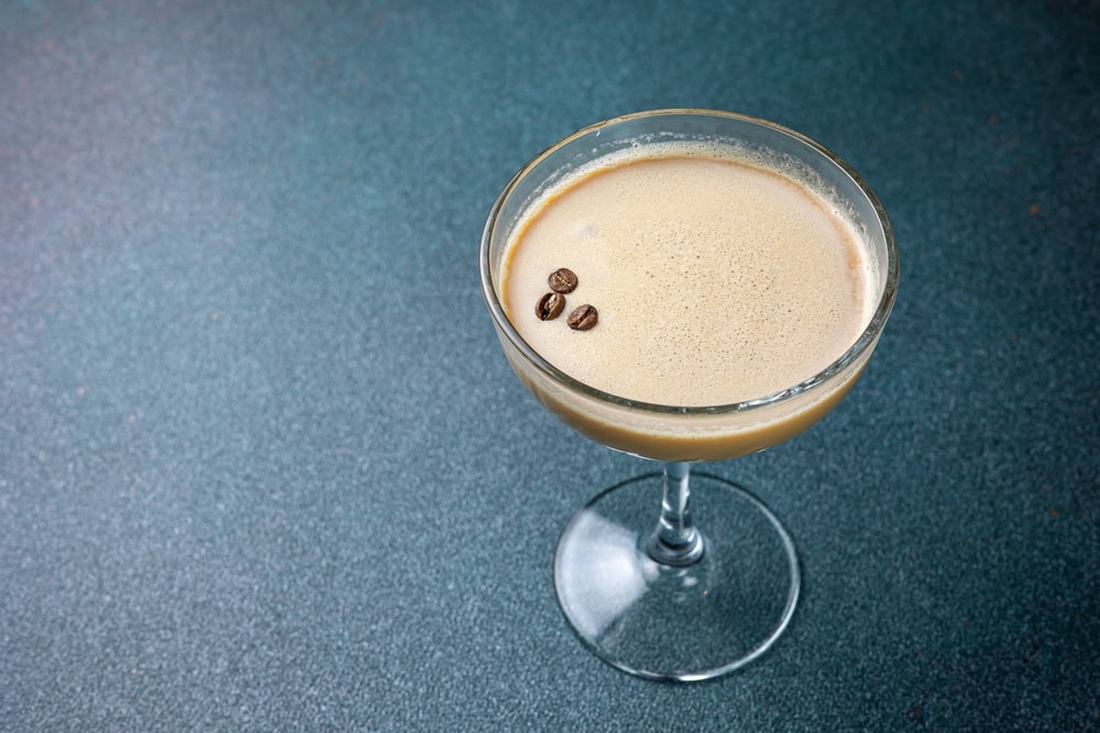 A coupe glass filled with a creamy light brown cocktail, garnished with three coffee beans, set on a textured blue surface.