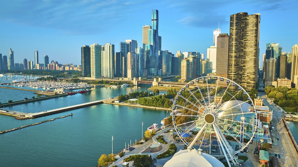 A large Ferris wheel stands near the waterfront with a marina and tall skyscrapers of a city skyline in the background under a clear blue sky.