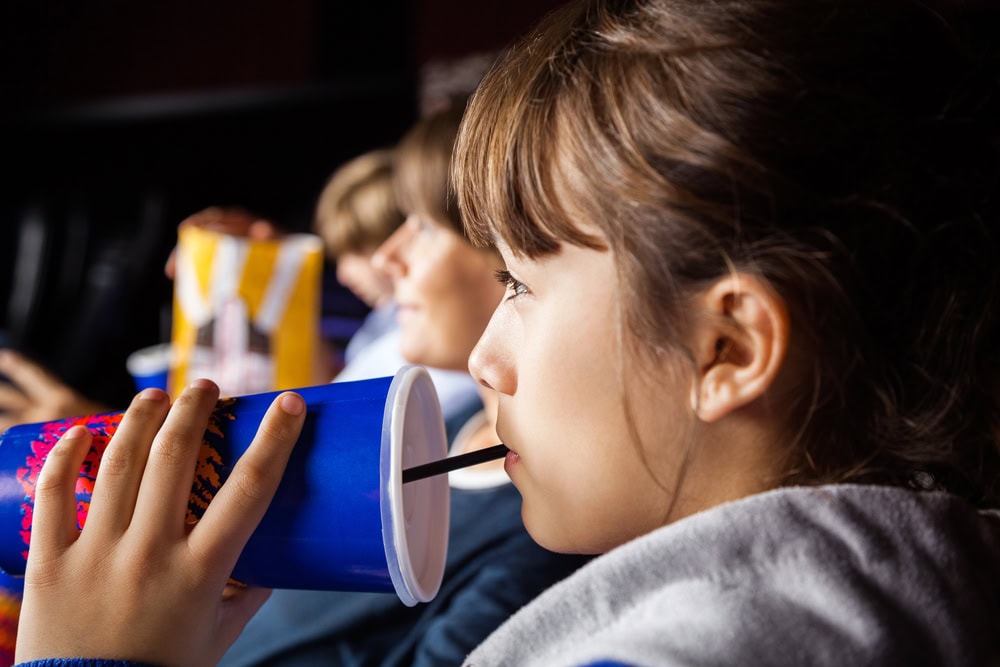 A young person sits in a movie theater, drinking from a large blue cup with a straw, with others and popcorn buckets in the background.