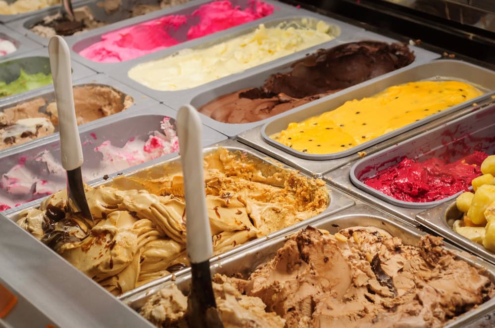 Assorted tubs of colorful ice cream and gelato on display in a shop, with serving scoops placed in some of the containers.