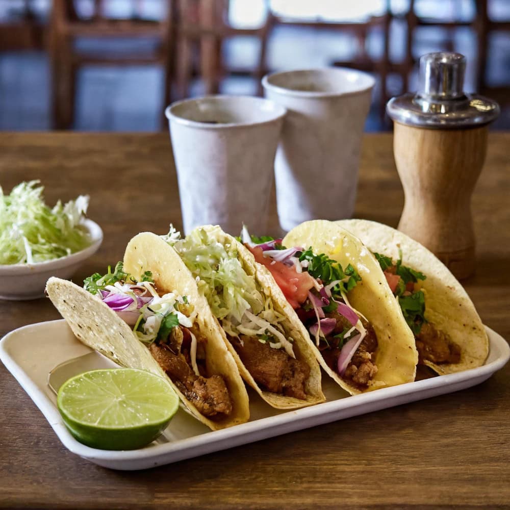Three tacos filled with fried fish, lettuce, tomato, onions, and cilantro on a tray with a halved lime; cups, lettuce, and a pepper grinder in the background.