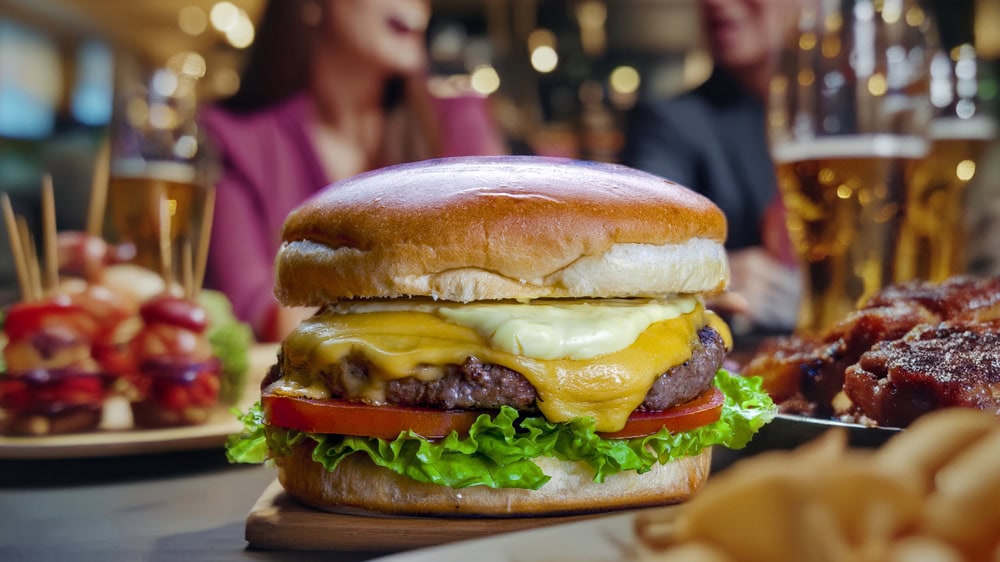 Close-up of a cheeseburger with lettuce, tomato, and melted cheese on a wooden board, with blurred people, appetizers, and drinks in the background.