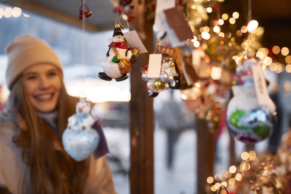 A woman in a beige hat smiles at a holiday market stall displaying colorful glass ornaments and festive lights.