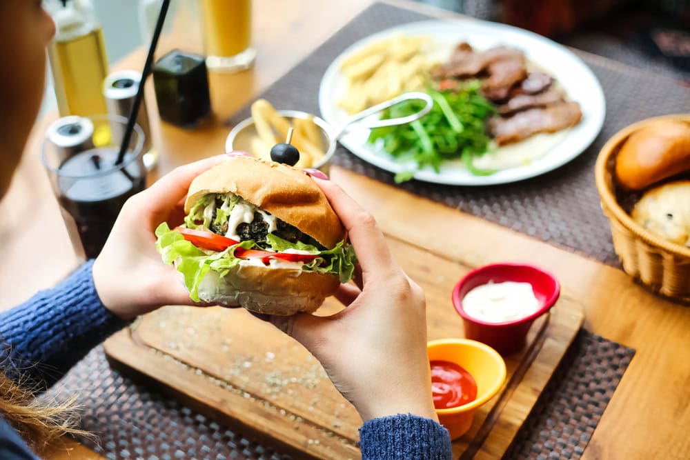 A person is holding a hamburger with lettuce, tomato, cheese, and meat. Sauces and a drink are nearby, with a plate of steak, fries, and salad in the background.