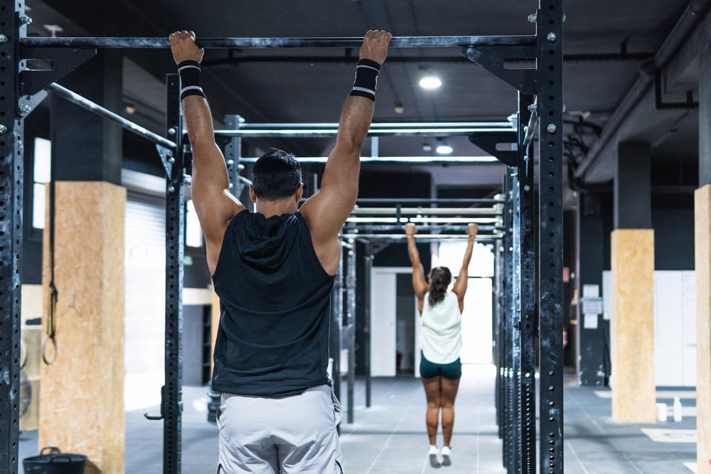 Two people hanging from pull-up bars in a gym, facing away from the camera. The gym has dark walls and fitness equipment in the background.