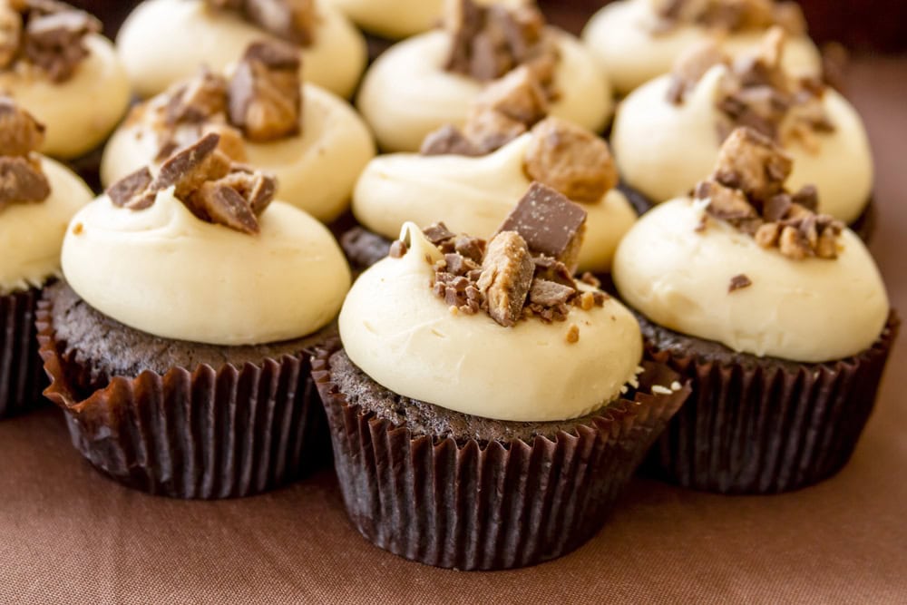 Chocolate cupcakes with light-colored frosting, topped with chopped chocolate and candy pieces, arranged closely together on a brown surface.