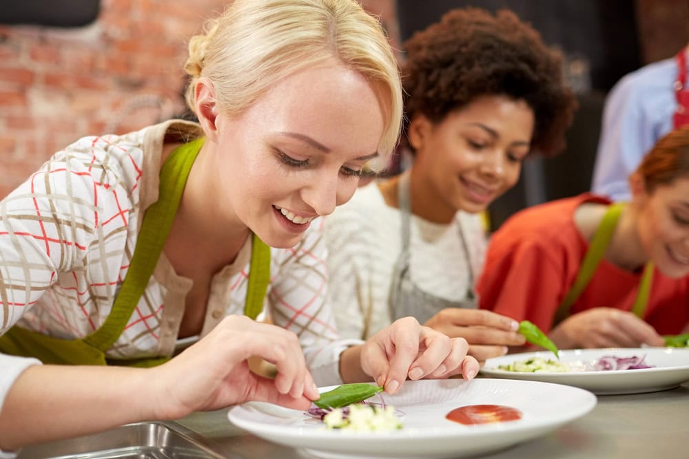 Three people wearing aprons carefully plating food on white dishes at a kitchen counter.