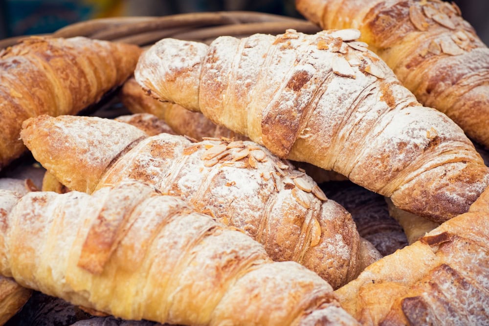 A close-up of several golden brown croissants dusted with powdered sugar and almond slices, stacked in a basket.