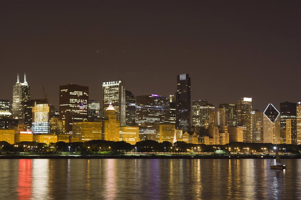 Chicago skyline at night with illuminated buildings reflected in the calm water, seen from across Lake Michigan.
