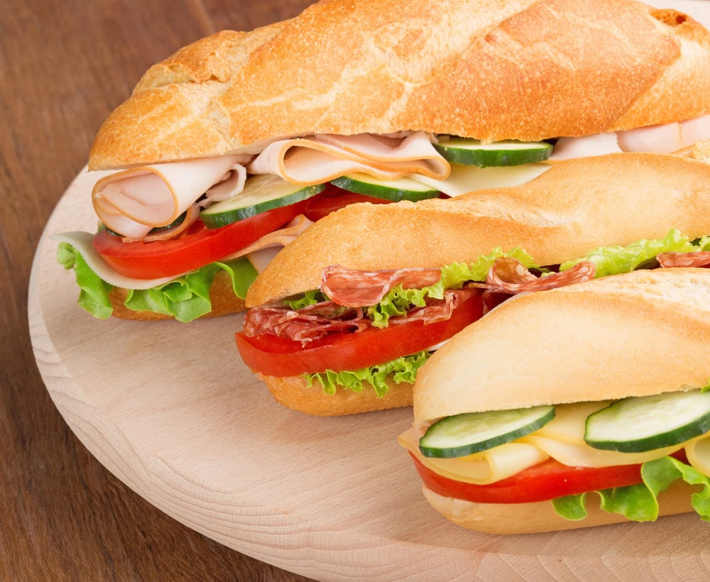 Three baguette sandwiches with lettuce, tomato, cucumbers, cheese, and sliced deli meats are arranged on a wooden board.