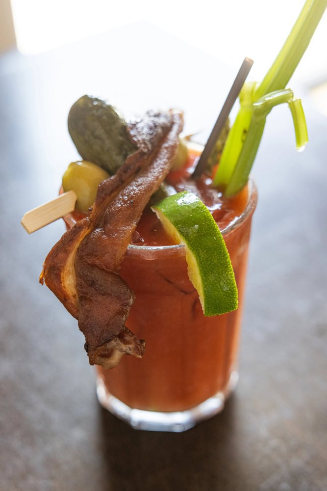 A Bloody Mary cocktail garnished with bacon, a lime wedge, celery stalk, pickle, and olives on a wooden skewer.