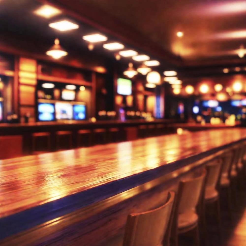 A long, empty wooden bar counter with chairs lined up, warm lighting, and multiple TV screens in the background.