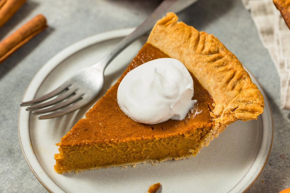 A slice of pumpkin pie with a dollop of whipped cream on a white plate, next to a fork.