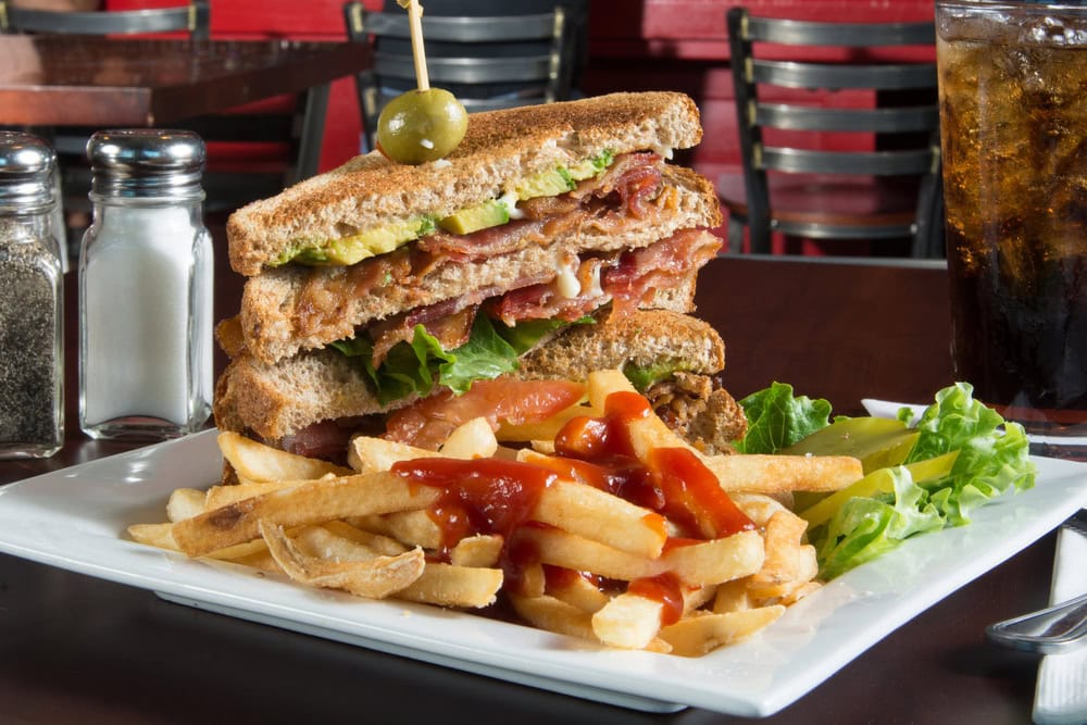 A club sandwich with lettuce, tomato, bacon, and avocado is stacked on wheat bread, served with a side of French fries topped with ketchup, lettuce garnish, and a glass of soda.