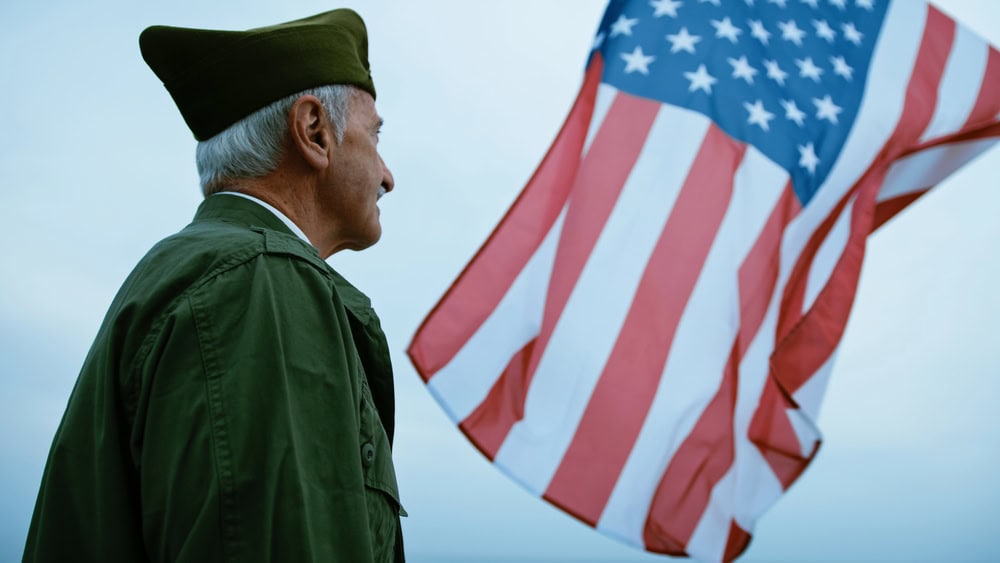 An older man in military attire stands outdoors, looking at a large American flag waving in the wind.