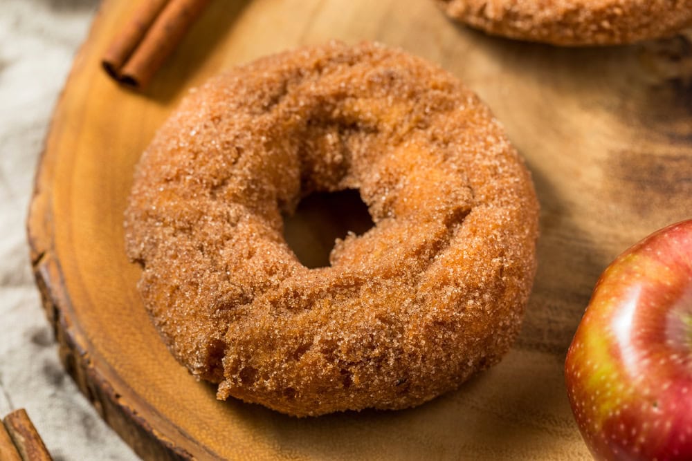 A cinnamon sugar-coated apple cider donut sits on a wooden surface next to an apple and a cinnamon stick.