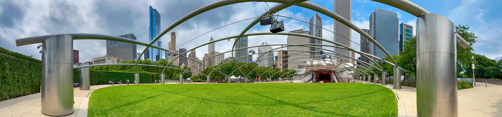 A panoramic view of an outdoor amphitheater with curved metal beams, a green lawn, and tall city buildings in the background under a partly cloudy sky.