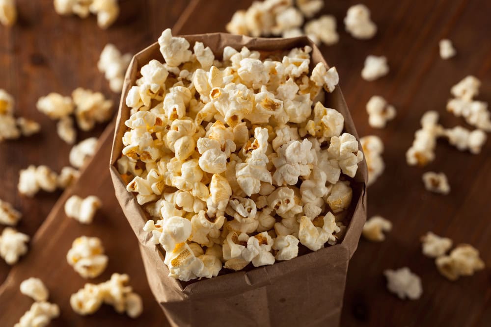 A brown paper bag filled with popped popcorn sits on a wooden surface, with some popcorn scattered around it.