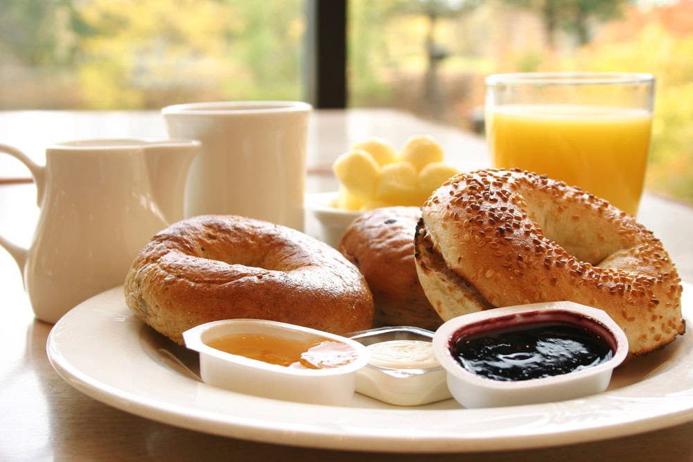 A plate with two bagels, small containers of jam, cream cheese, and butter, two cups, a pitcher, and a glass of orange juice on a table.