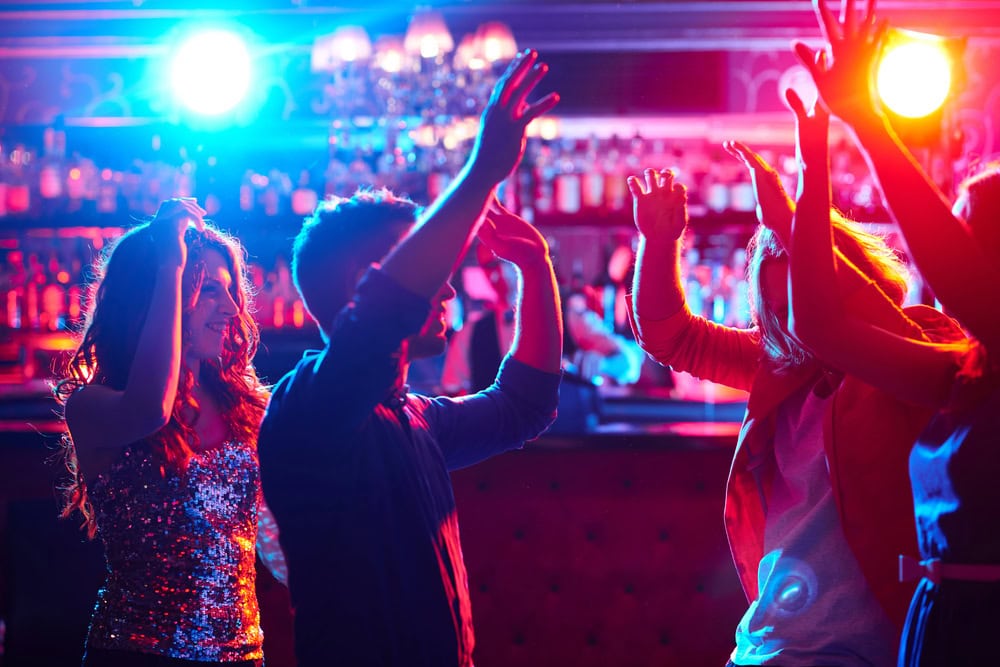 Group of people dancing in a dimly lit nightclub with colorful lights and a bar in the background.
