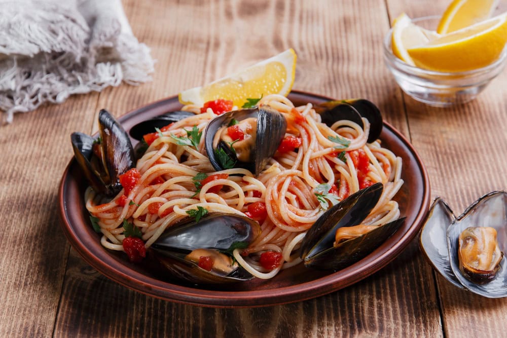 A plate of spaghetti with tomato sauce and mussels, garnished with parsley, served with lemon wedges on a wooden table.