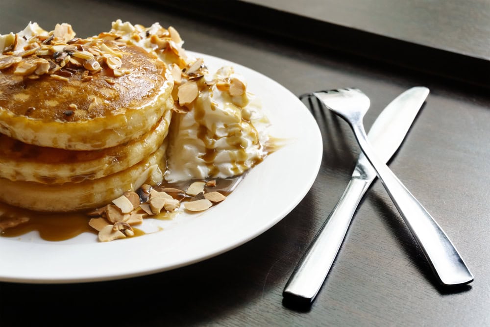 A stack of pancakes topped with syrup and sliced almonds sits next to a dollop of whipped cream on a white plate, with a fork and knife placed beside it.