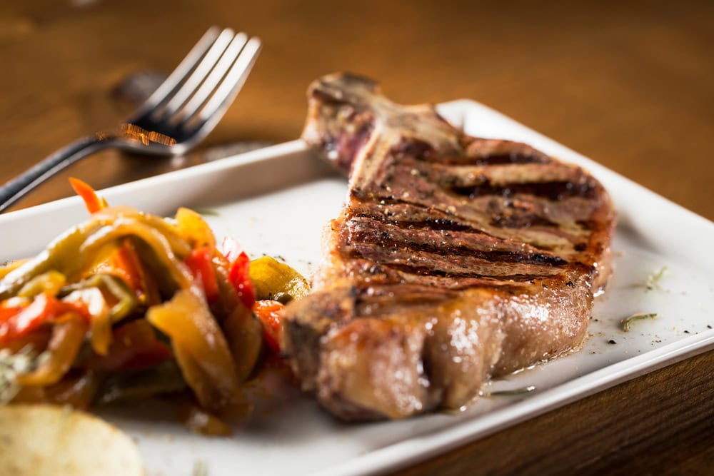 A grilled steak with visible char marks served on a white plate next to sautéed bell peppers, with a fork in the background.