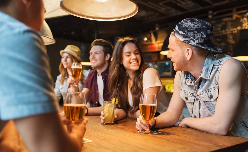 Five young adults sit at a bar counter, talking and holding drinks. The atmosphere is casual and relaxed, with warm lighting and a blurred background.