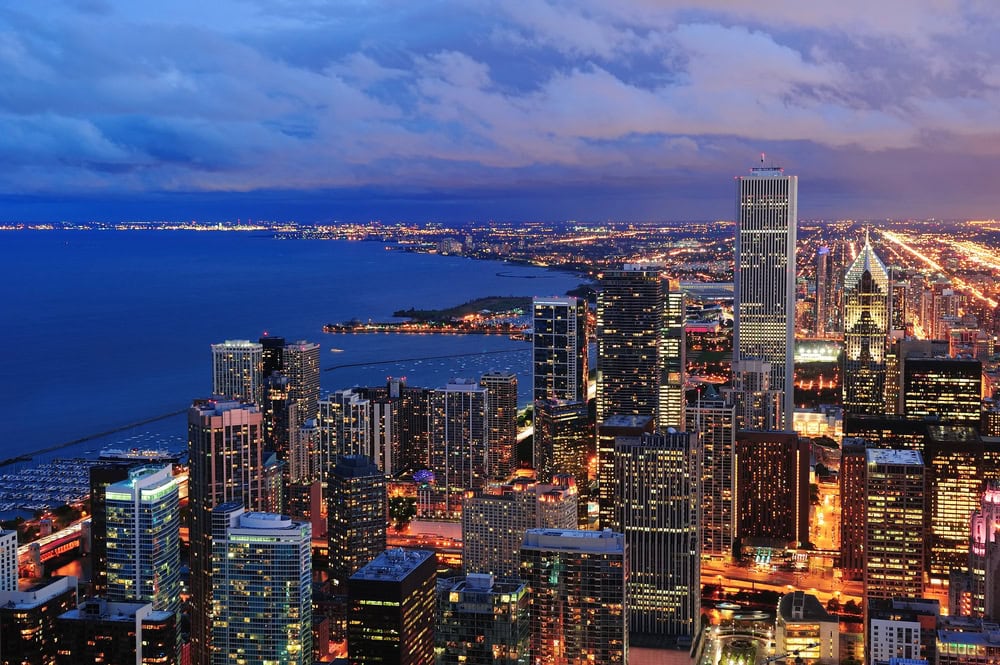 Aerial view of a city skyline at dusk with illuminated buildings, a large lake on the left, and streets stretching into the distance under a partly cloudy sky.