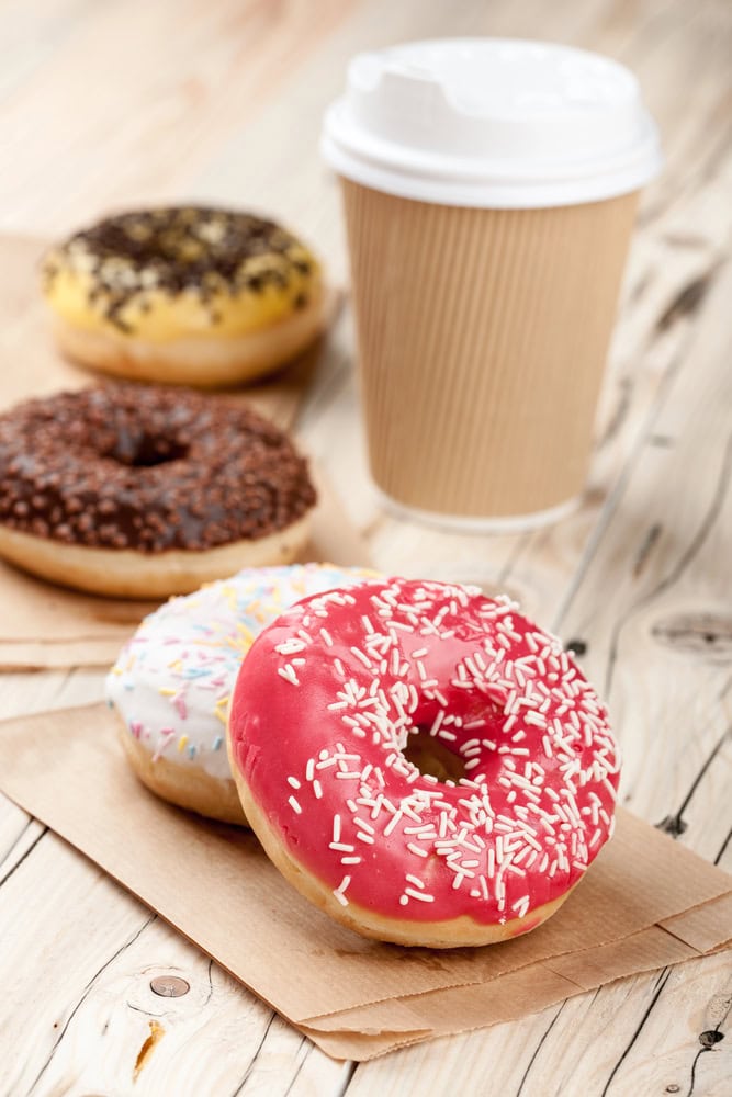 A pink glazed donut with sprinkles in front of three other donuts and a takeaway coffee cup on a wooden surface.