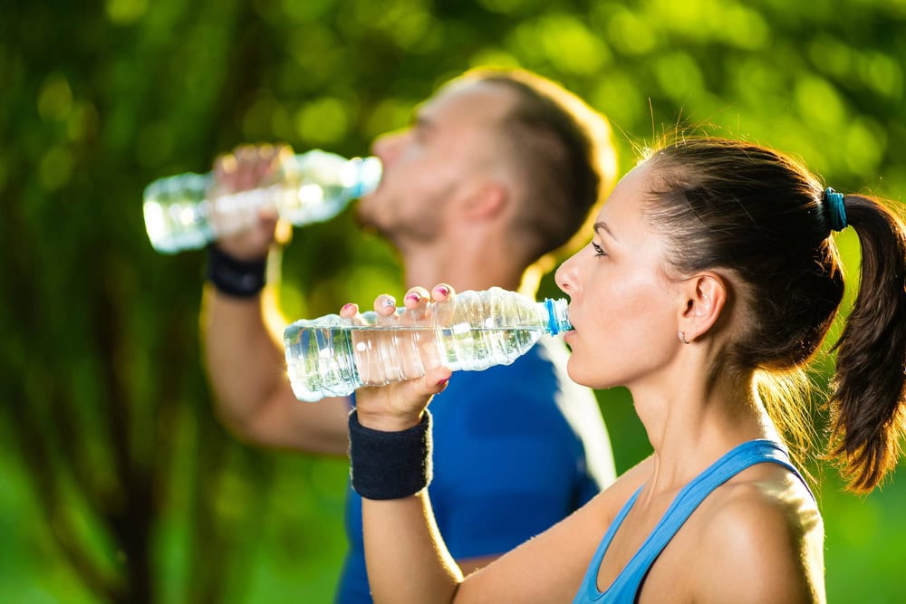 A man and a woman in athletic clothing drink water from plastic bottles outdoors, with greenery in the background.